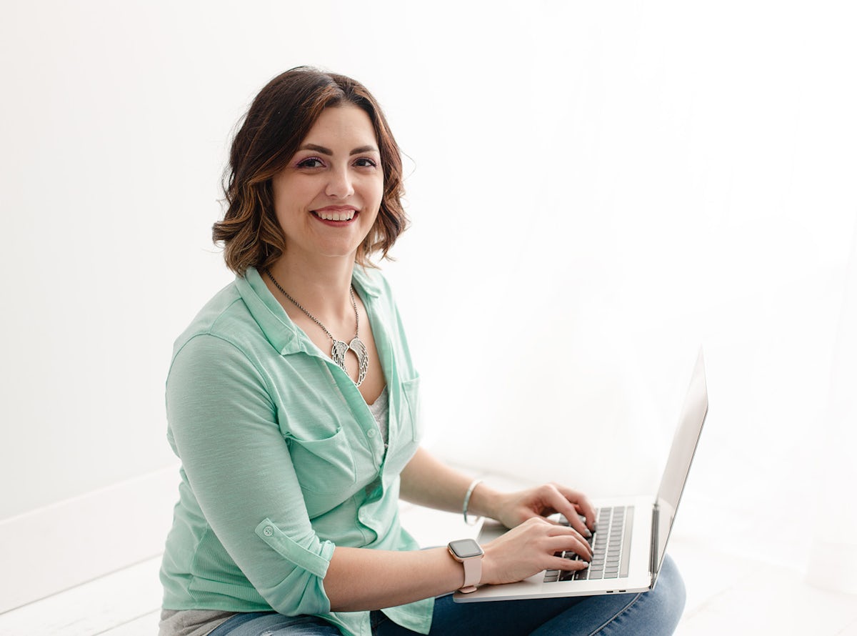 Smiling white woman wearing a mint green button up blouse and seated cross-legged on the floor, looking into the camera and holding an open Macbook Pro on her lap